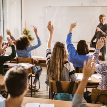 Rear view of large group of students raising their hands to answer the question on a class.